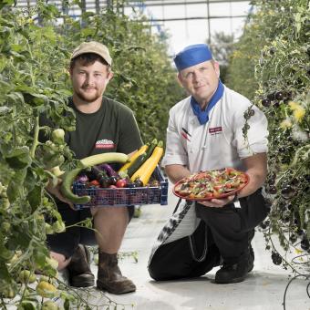 Eden gardener and chef kneeling down in greenhouse between rows of tomato plants, holding out freshly picked produce